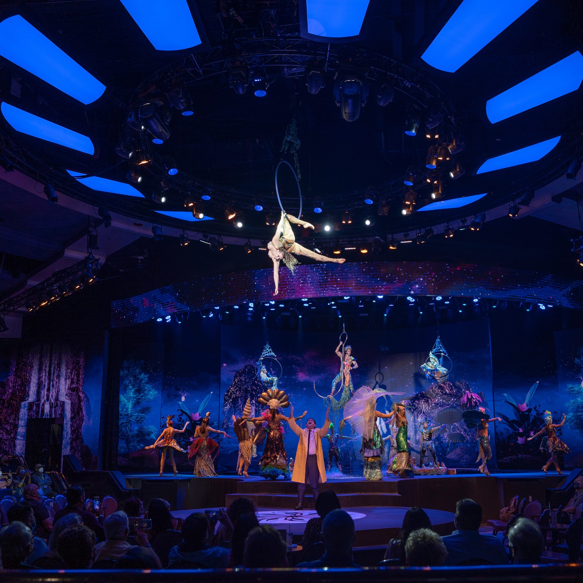 Acrobat performing aerial hoop act surrounded by dancers in colorful costumes at WOW Las Vegas